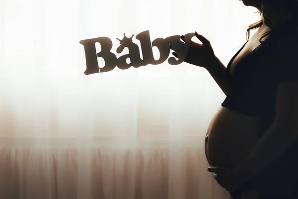 창 앞에 서 있는, 배꼽을 들고 임신한 여자의 실루엣. 임신한 여자 아기 편지를 들고 — 스톡 사진