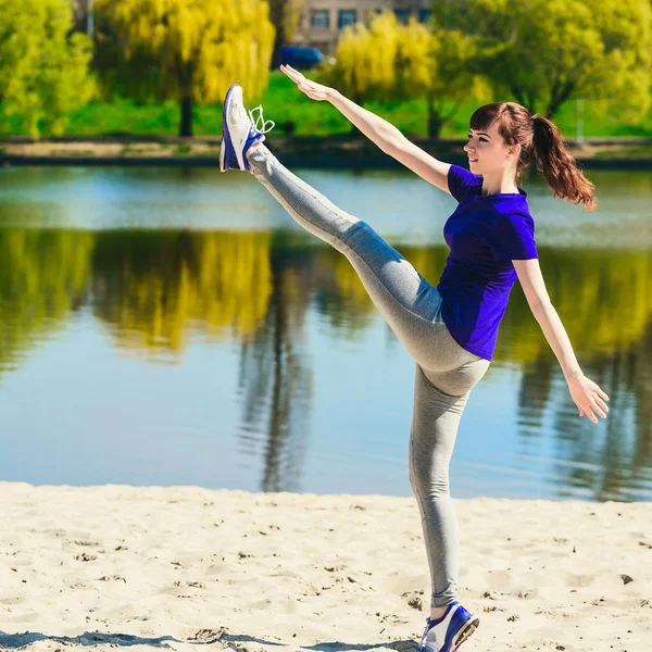 La giovane donna allunga la gamba. Indossa abiti sportivi blu. La spiaggia e il fiume sono sullo sfondo . — Foto Stock