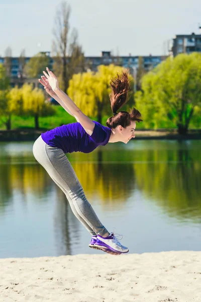 Joven morena en una camisa deportiva azul haciendo un salto en la playa. Mujer haciendo ejercicio de fitness al aire libre . — Foto de Stock