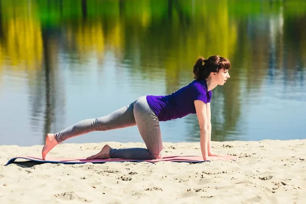 brunette in blue sports shirt on fitness mat doing exercises on beach. Woman doing fitness exercise outdoors