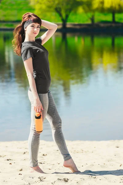 Meisje in de sport kleding houdt een fles water, weg op zoek en glimlachend, staande op het strand na training — Stockfoto