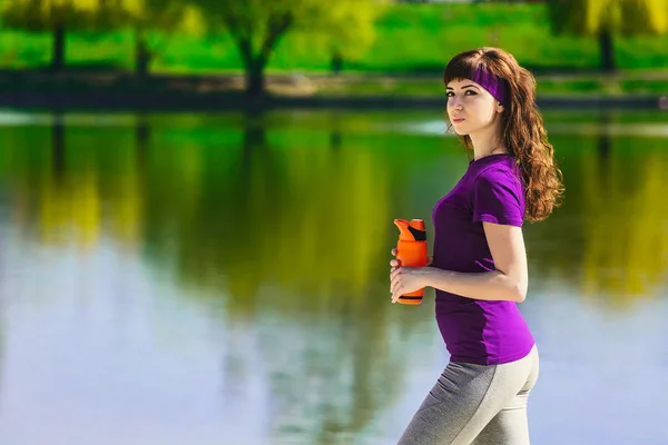在运动服的女孩拿着一瓶水，视线和微笑，站在沙滩上后，锻炼 — 图库照片