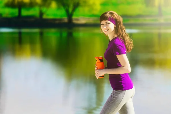 Ragazza in abiti sportivi sta tenendo una bottiglia d'acqua, guardando altrove e sorridendo, in piedi sulla spiaggia dopo l'allenamento — Foto Stock