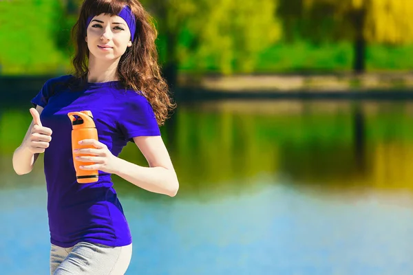 women in sport clothes is holding a bottle of water, looking away and smiling, standing on the beach after workout