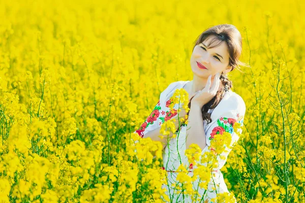 Belle jeune femme en ukrainien brodé debout dans un champ de fleurs de viol jaune — Photo