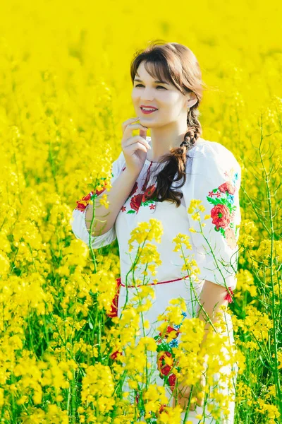 Mulher bonita em ucraniano bordado em pé em um campo de flores de estupro amarelo — Fotografia de Stock