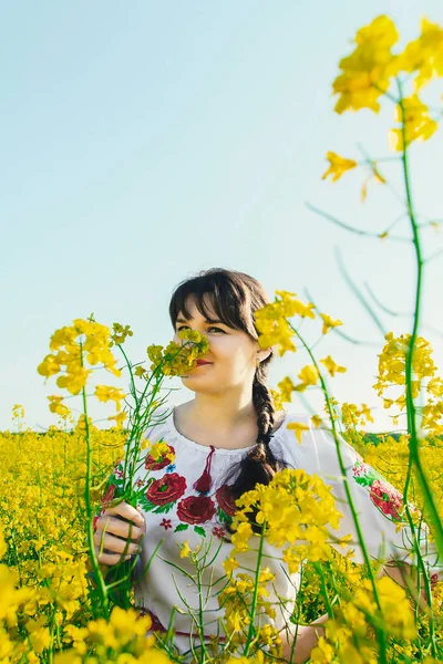 Belle jeune femme en ukrainien brodé debout dans un champ de fleurs de viol jaune — Photo