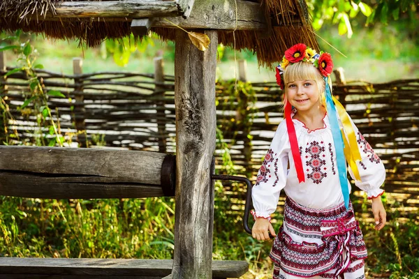 A little girl in a Ukrainian folk costume stands near an old wooden kolodiaz. A girl takes water from a well — Stock Photo, Image