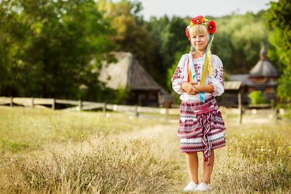 Bambina in costume popolare ucraino si trova sul campo su uno sfondo di vecchie capanne di legno — Foto Stock