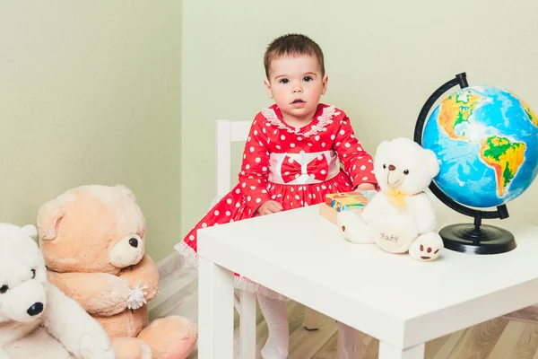 Ein einjähriges Mädchen in rotem Kleid sitzt an einem Tisch mit einem Buch, einer Weltkugel und einem Teddybär. — Stockfoto