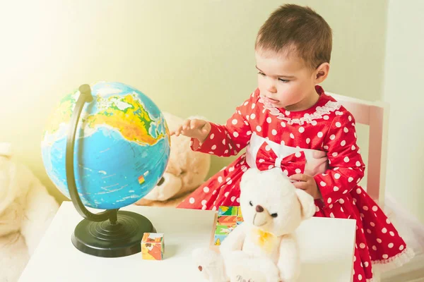 Ein einjähriges Mädchen in rotem Kleid sitzt an einem Tisch mit einem Buch, einer Weltkugel und einem Teddybär. — Stockfoto