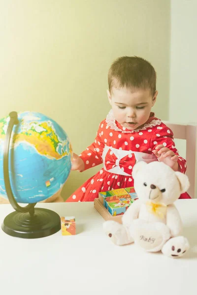 Ein einjähriges Mädchen in rotem Kleid sitzt an einem Tisch mit einem Buch, einer Weltkugel und einem Teddybär. — Stockfoto