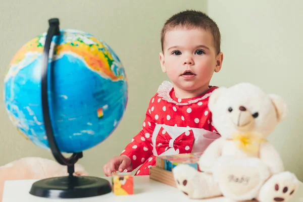 Ein einjähriges Mädchen in rotem Kleid sitzt an einem Tisch mit einem Buch, einer Weltkugel und einem Teddybär. — Stockfoto