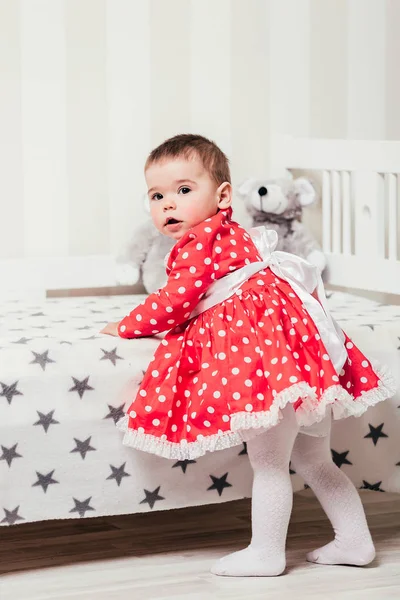 A one-year-old girl in a red dress takes her first steps holding onto the bed in the room — Stock Photo, Image