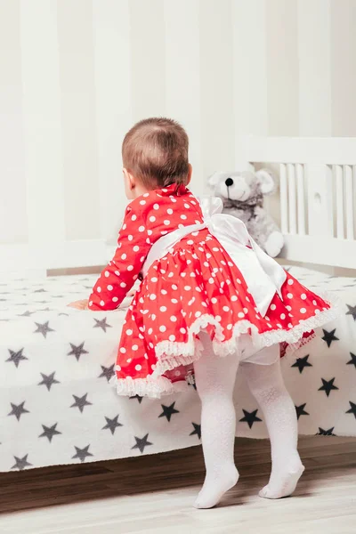 Ein einjähriges Mädchen im roten Kleid macht ihre ersten Schritte, indem es sich am Bett im Zimmer festhält — Stockfoto