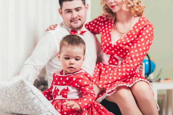 A one-year-old girl in a red dress sits on a bed in a room with her parents — Stock Photo, Image
