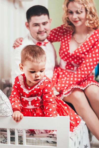 Una niña de un año con un vestido rojo se sienta en una cama en una habitación con sus padres —  Fotos de Stock