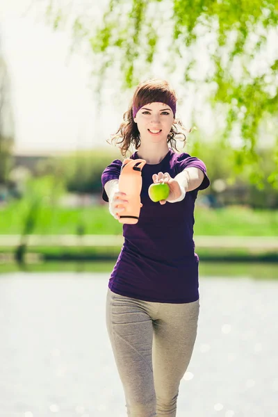 Giovane atleta donna carina in un giubbotto blu con una bottiglia d'acqua e una mela e guardando la fotocamera — Foto Stock