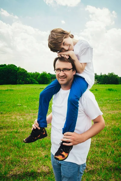 Der kleine Junge und sein hübscher junger Vater lächeln, während sie sich im Park ausruhen. Sohn sitzt auf den Schultern seines Vaters. — Stockfoto