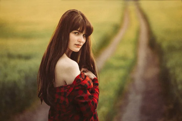 Attractive young brunette woman stands in a field on a dirt road in a red checkered shirt.
