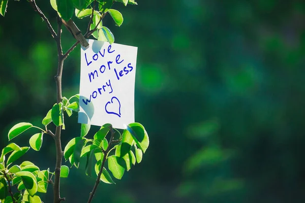 Motivating phrase love more worry less. On a green background on a branch is a white paper with a motivating phrase