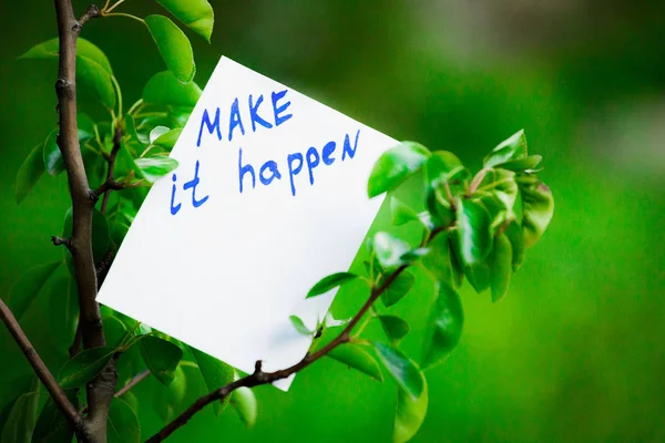 Motivating phrase make it happen. On a green background on a branch is a white paper with a motivating phrase.