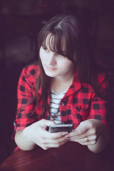 Young brunette woman in red checkered shirt sitting near window with phone in her hands — Stock Photo, Image