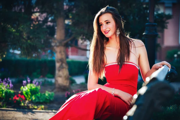 Krásná mladá brunetka žena na sobě červené šaty, chůzi na ulici. Štíhlá dívka v červené dlouhé šaty sedí na lavičce v parku — Stock fotografie