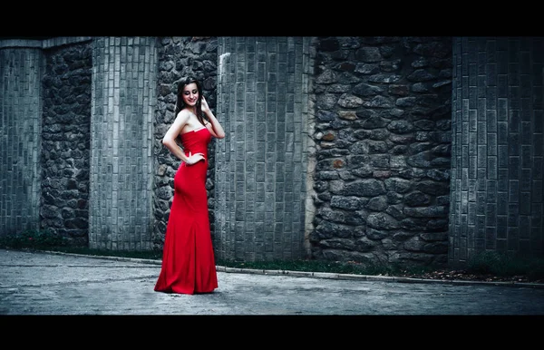 Ragazza snella in rosso abito lungo si erge su sfondo muro di pietra. foto di moda — Foto Stock