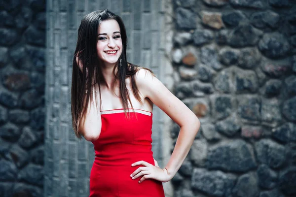 Chica delgada en rojo vestido largo se encuentra en la pared de piedra de fondo. —  Fotos de Stock