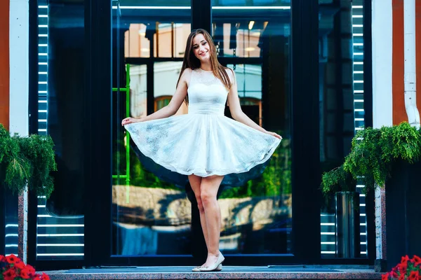 Mujer joven en un vestido blanco corto está de pie sosteniendo un vestido en las manos en el fondo de una puerta de cristal —  Fotos de Stock