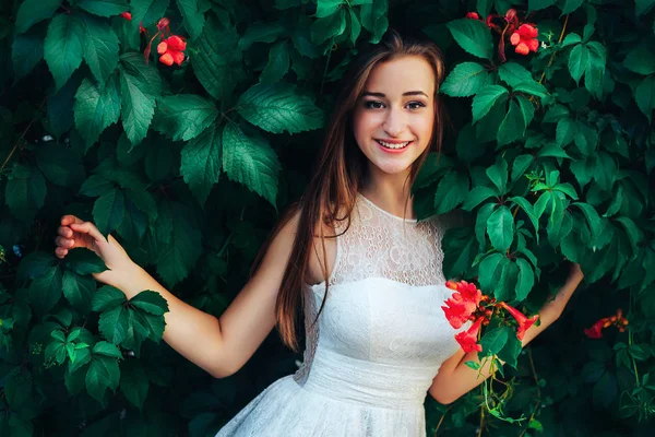 Joven mujer morena hermosa en vestido blanco se encuentra en el fondo de la pared con hojas verdes y flores . —  Fotos de Stock