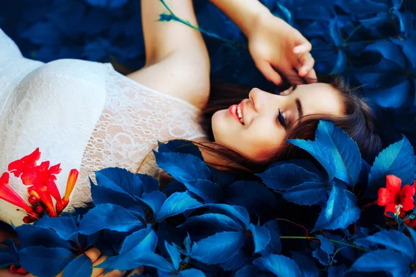 Schöne junge brünette Frau in weißem Kleid liegt auf fantastischen blauen Blättern und Blüten.fantastische Landschaft. — Stockfoto