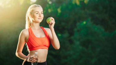 Kırmızı bir üst ve siyah pantolon içinde genç bir sarışın bir şişe su ve bir yeşil elma açık havada tutar.
