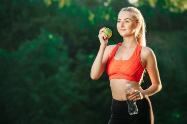 Kırmızı bir üst ve siyah pantolon içinde genç bir sarışın bir şişe su ve bir yeşil elma açık havada tutar.