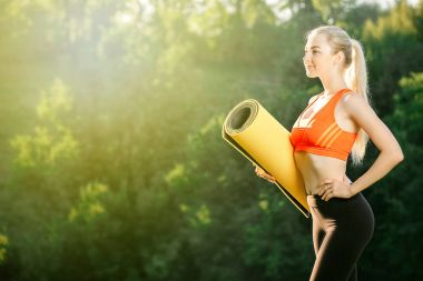 Kırmızı bir üst ve siyah pantolon içinde genç bir sarışın bir mat bir çeşme doğa için tutar. Bir kadın jimnastik için hazırlar