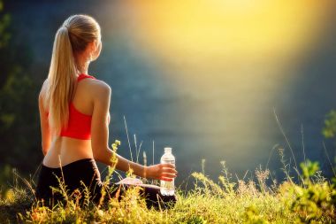 Genç sarışın bir kırmızı üst ve siyah pantolon içinde doğa cephede oturur. Sportif bir kadın bir şişe su tutuyor.