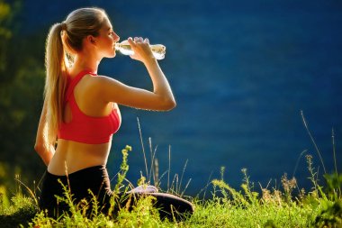 Genç sarışın bir kırmızı üst ve siyah pantolon içinde doğa cephede oturur. Sportif bir kadın bir şişe su tutuyor.