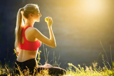 Genç sarışın bir kırmızı üst ve siyah pantolon içinde doğa cephede oturur. Sportif bir kadın bir yeşil elma onun elinde tutar.