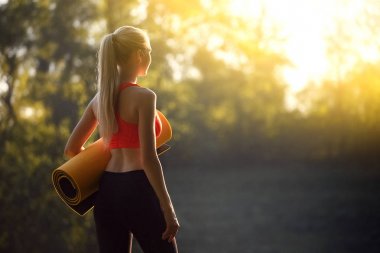 Kırmızı bir üst ve siyah pantolon içinde genç bir sarışın bir mat bir çeşme doğa için tutar. Bir kadın jimnastik için hazırlar