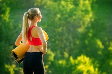 Kırmızı bir üst ve siyah pantolon içinde genç bir sarışın bir mat bir çeşme doğa için tutar. Bir kadın jimnastik için hazırlar