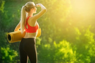 Kırmızı bir üst ve siyah pantolon içinde genç bir sarışın bir mat bir çeşme doğa için tutar. Bir kadın jimnastik için hazırlar