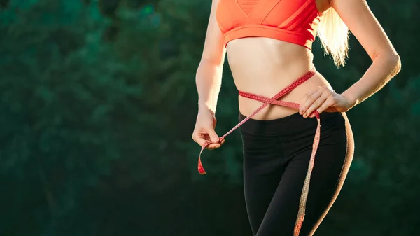 Een jonge blonde in een rode top en zwarte broek meet haar taille met een liniaal lint in de natuur. — Stockfoto