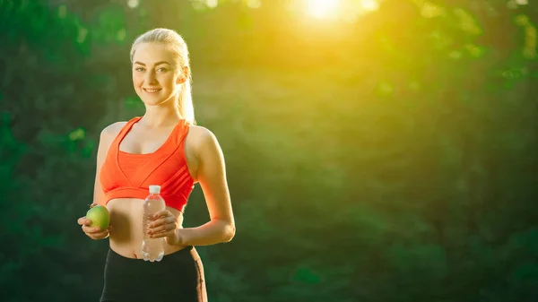 A young blonde in a red top and black pants holds a bottle of water and a green apple in the open air. — Stock Photo, Image