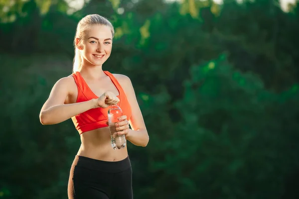 Una giovane bionda con un top rosso e pantaloni neri tiene in mano una bottiglia d'acqua e la apre a bere in natura . — Foto Stock