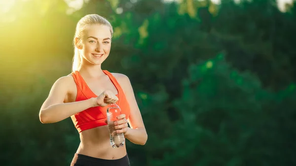 Una giovane bionda con un top rosso e pantaloni neri tiene in mano una bottiglia d'acqua e la apre a bere in natura . — Foto Stock