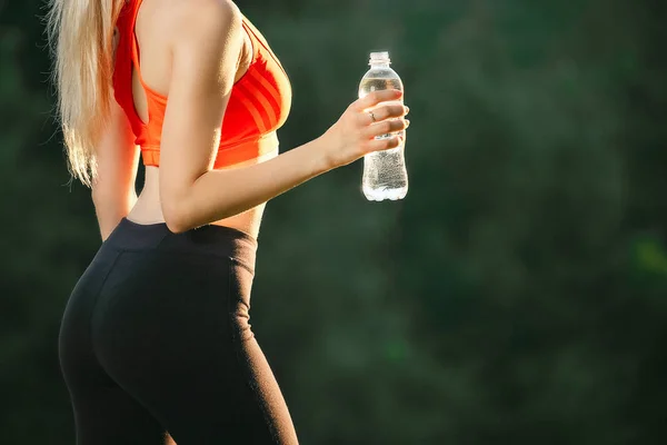 Una giovane bionda con un top rosso e pantaloni neri tiene in mano una bottiglia d'acqua. Concetto di alimentazione sana e sport — Foto Stock