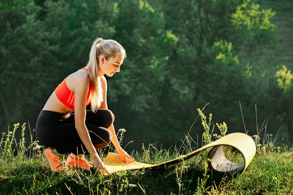 Une jeune blonde en haut rouge et pantalon noir tient un tapis pour une fontaine dans la nature. Une femme se prépare à la gymnastique — Photo