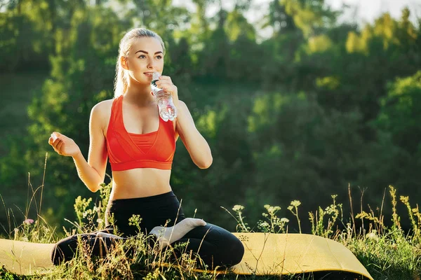 Giovane bionda in top rosso e pantaloni neri si siede con una bottiglia d'acqua su un tappeto giallo per il fitness in natura . — Foto Stock