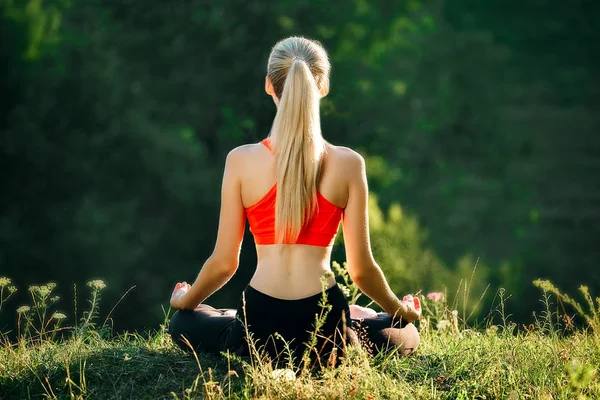 Egy fiatal szőke egy piros tetején ül a fűben, a fitness a természetben. A sportoló előkészíti a torna számára. — Stock Fotó